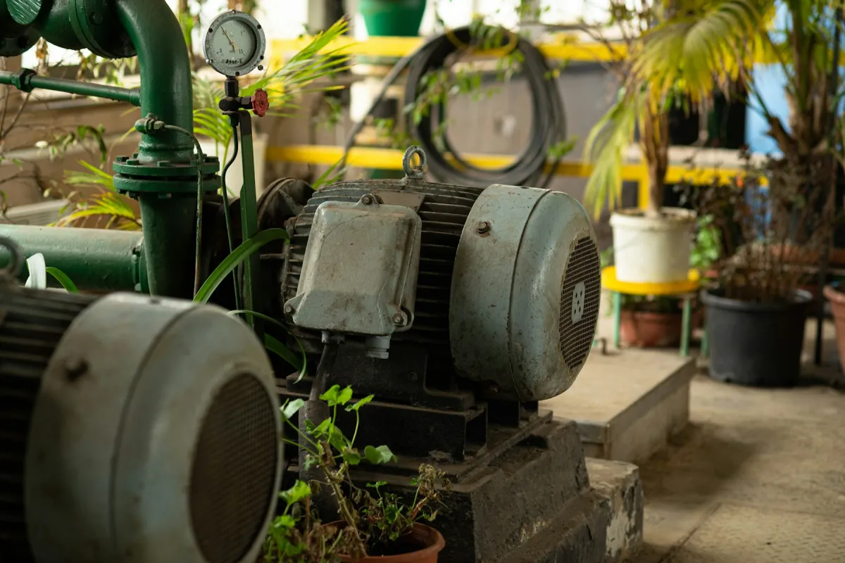 Motor dan pompa di atas base frame dengan pressure gauge pada pipa keluaran, dikelilingi tanaman di area utilitas