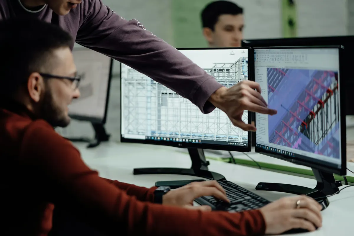 Para engineer bekerja di meja, salah satunya menunjuk model CAD 3D sebuah struktur di layar monitor