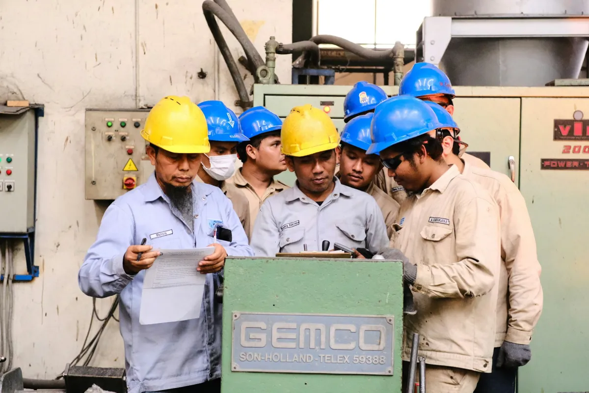 Sekelompok teknisi maintenance berhelm kuning dan biru berkumpul di depan panel kontrol mesin sambil membahas sebuah dokumen