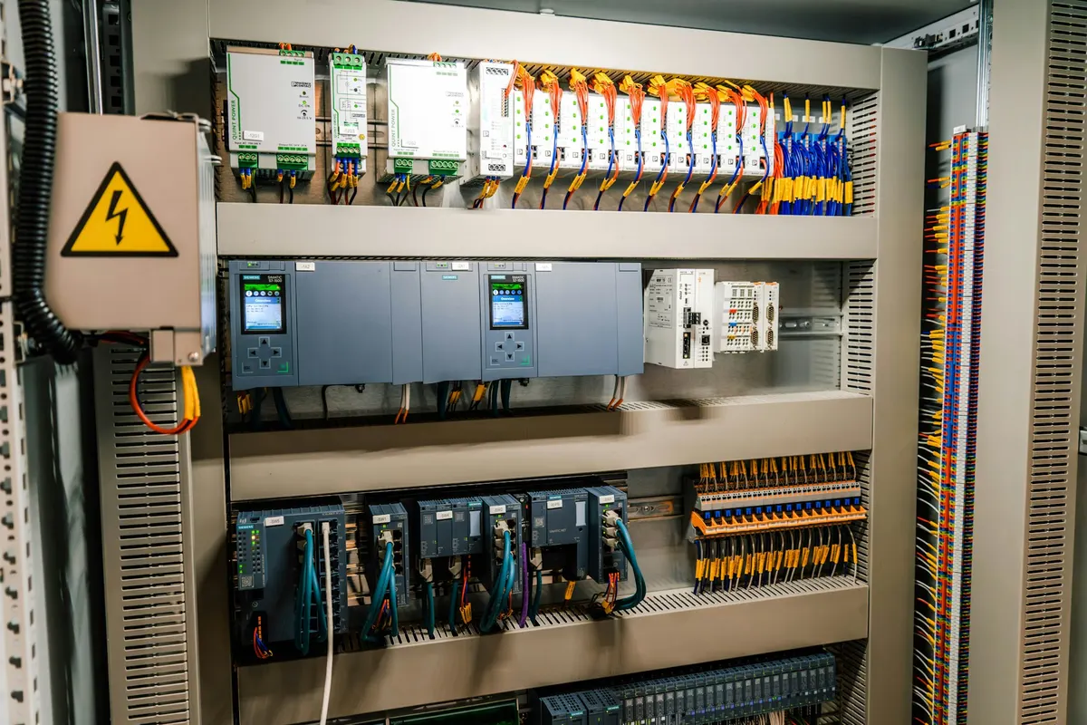 Panel kontrol listrik industri yang terbuka dengan deretan breaker, terminal block, dan tanda peringatan tegangan tinggi berwarna kuning